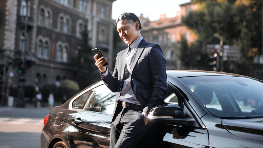 Executive Transfer – Stay Connected While You Travel Businessman leaning on a black car while checking his phone in the city